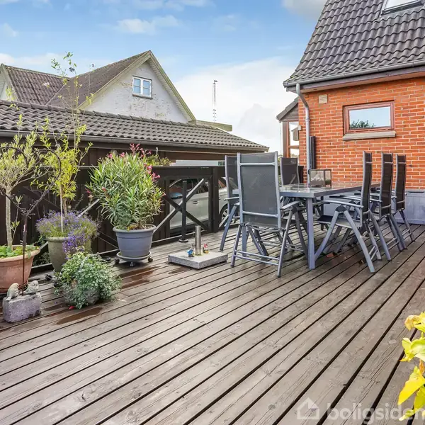 Terrasse med et sæt sorte haveborde og stole. Der er mange blomster og planter i potter omkring. Baggrunden viser et rødt murstenshus og et nærliggende tag.