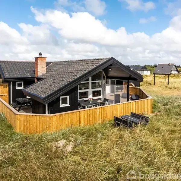 Træhus med sort tag og store hvide vinduer står på en sandet, græsbevokset grund omgivet af lavt hegn. Tilhørende udendørsmøbler er placeret på terrassen.