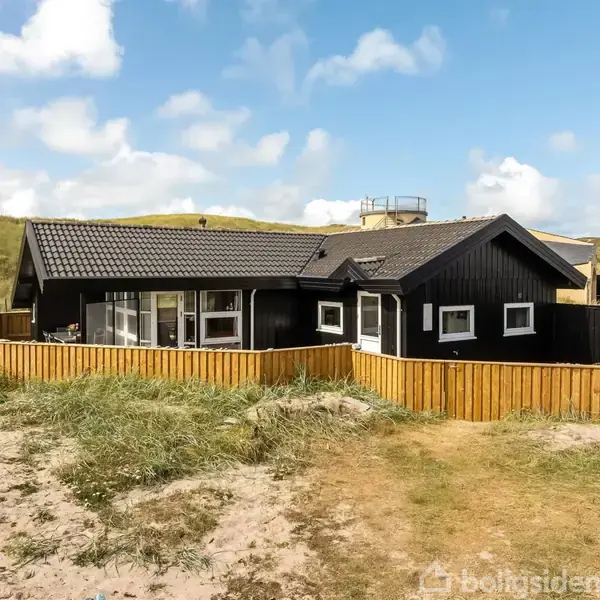 Sort træhus står stille omgivet af sand og klitter. Huset har en veranda med glaspaneler og et skråt tag. Græs vokser i forgrunden, mens himlen er let skyet.