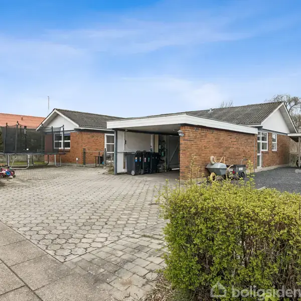 En murstensbungalow med garage er omgivet af flisebelagt forplads og grønt buskads. En trampolin står ved siden af huset i en forstadshave. Tørt og solrigt vejr.