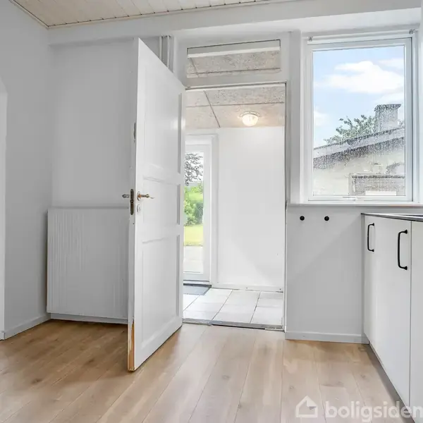Dør åbner mod en lys, moderne køkkenindgang med hvide skabe og flisegulv, set fra trælaminatgulvstue med buet åbning og radiator ved vindue til højre.
