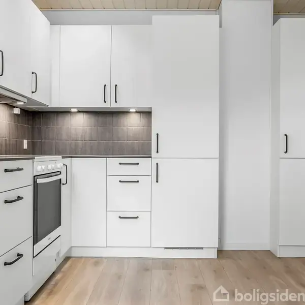 Køkken med hvide skabe og sorte håndtag, ovn placeret i midten, grå fliser over bordplade, lyse trægulve og spotlys under overskabe skaber et moderne look.