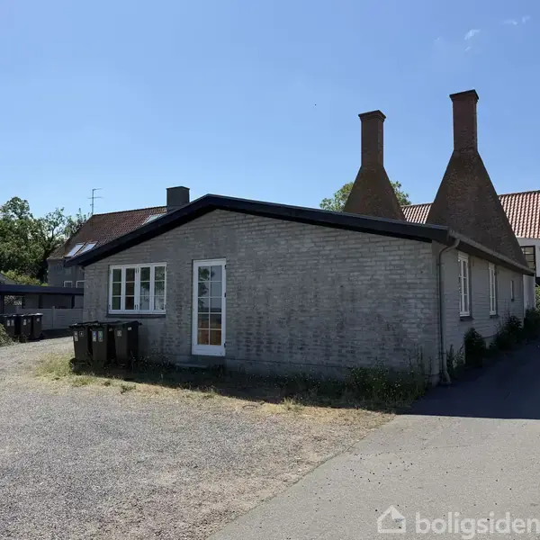 En grå murstensbygning med skråtag og to høje skorstene står i et solrigt, åbent område omgivet af andre huse og grønne træer. En grusvej løber langs bygningen.