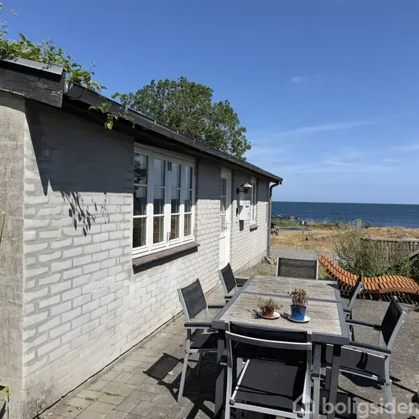 Et gråmalet murstenshus med hvide vinduer står ved siden af et bord med stole. Det er placeret på en terrasse med udsigt over havet og en blå himmel.