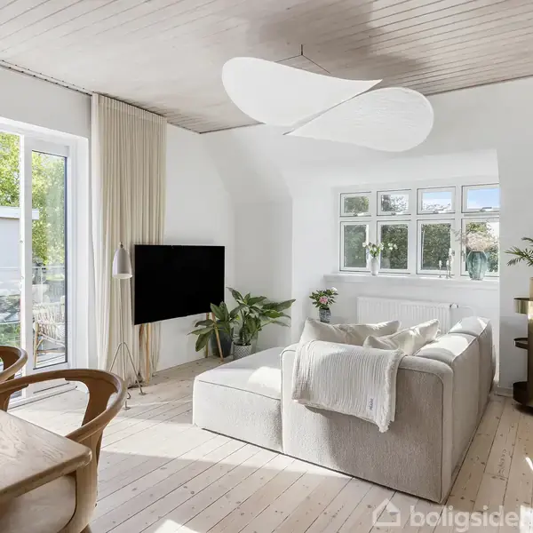 Lysegrå hjørnesofa i stue med trægulv, store vinduer og moderne belysning. Sofaen vender mod et fjernsyn på venstre side. Rummet er lyst, med planter og hylder.