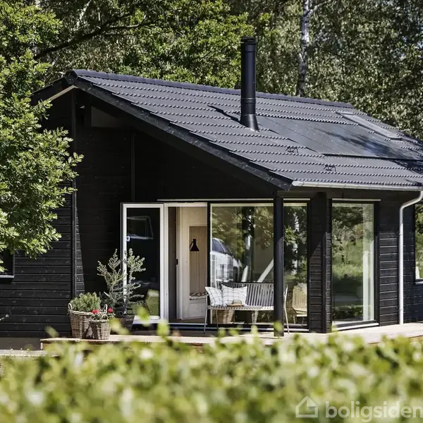 Et lille sort træhus med store vinduer står omgivet af grønne træer og buske. En terrasse med en hvid bænk ligger foran huset.