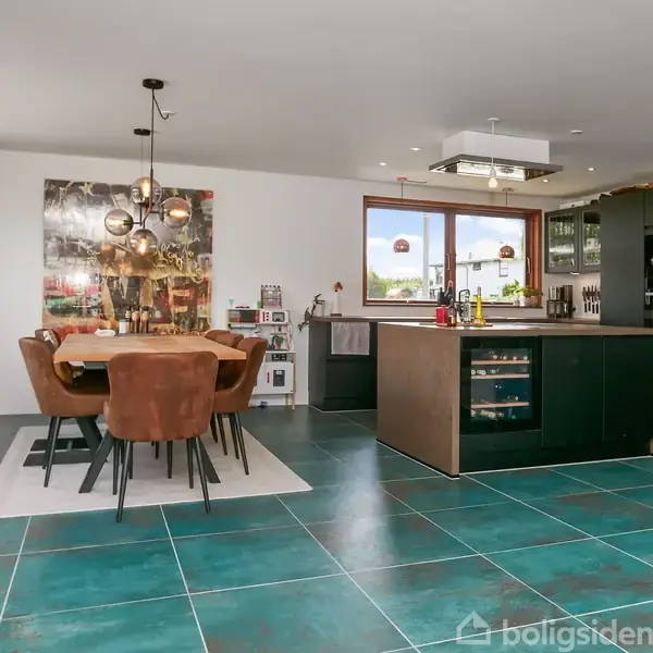 Et moderne køkkenalrum med store grønne fliser, spisebord med brune stole, og et køkkenområde med en køkkenø. Vægdekorationer og store vinduer med lysindfald præger rummet.
