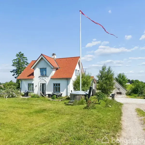 Et hvidt hus med rødt tag står på en stor grøn græsplæne under en blå himmel. En vindblæst flagstang står foran huset ved siden af en grusvej.