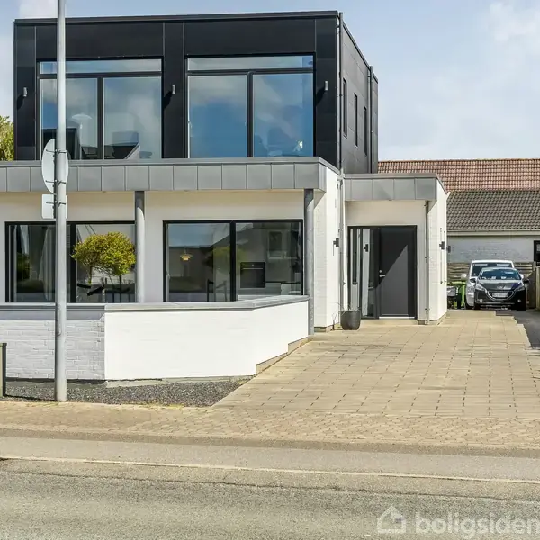 To-etagers moderne hus står stille ved en vej. Huset har store vinduer og fladt tag. Der er en indkørsel til højre med en bil parket ved garagen.