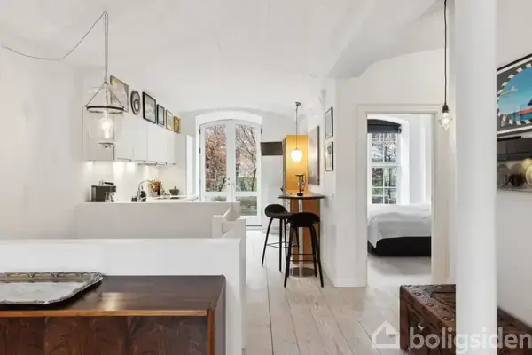 Stue med hvide vægge og lys trægulv, åbner op mod køkken med hvide skabe. To barstole står ved et lille bord. En dør fører til et soveværelse. En stor vinduesfront ses i baggrunden.