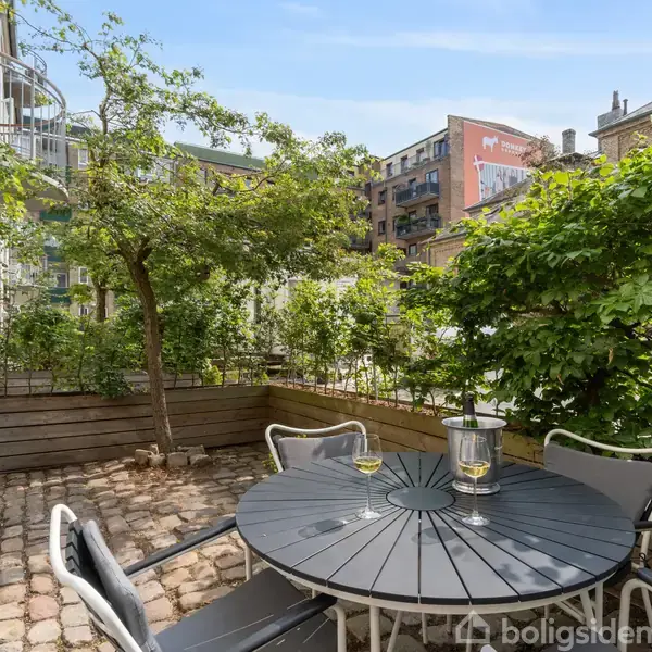 Rundt havebord med stole står på brosten i en gårdhave. Bordet har to vinglas og en isspand. Omkring bordet er grønt løv og bygninger i baggrunden.