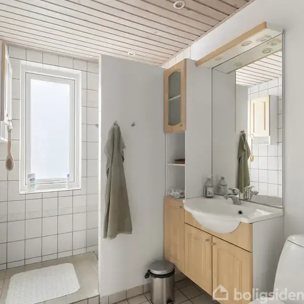 Badeværelse indeholder håndvask med spejl, bruser og toilet. Lyse fliser på vægge og gulv; naturligt lys kommer ind gennem et vindue. En håndklædeholder med håndklæde hænger ved siden af bruseren.