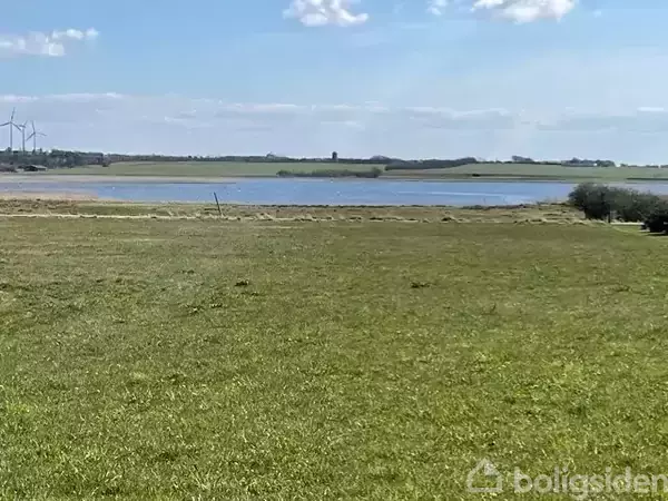 En grøn græsmark breder sig ud, mens en rolig sø ligger i baggrunden. Vindturbiner kan ses på horisonten under en solrig himmel med få skyer.