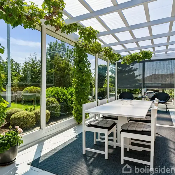 Et hvidt bord med fire stole står på en overdækket terrasse omgivet af glasvægge. Vinranker klatrer langs strukturen, og udenfor ses en velplejet have med buske og træer.