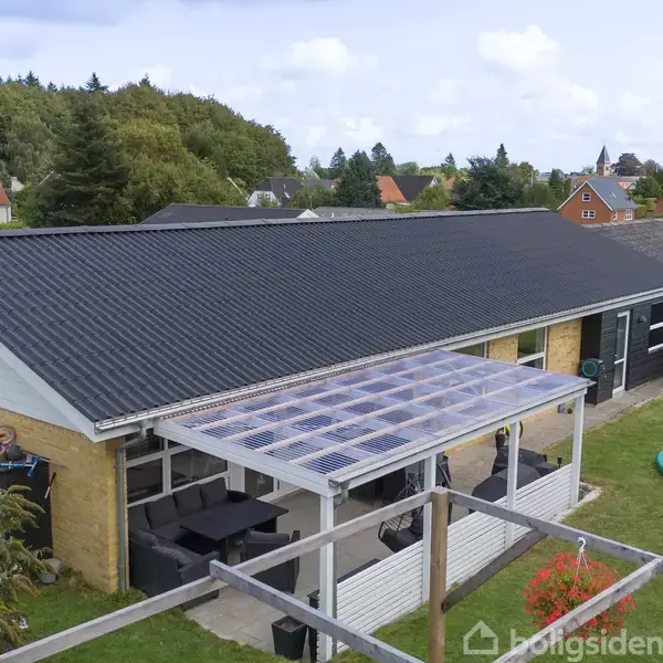 Et enfamiliehus med sort tag og gul murstensfacade. En overdækket terrasse med gennemsigtig tag ligger ved siden af huset. Haven har en trampolin og græsplæne.