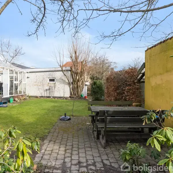 En have med grøn græsplæne og terrasse med et stort, sort havebord og bænke. Til venstre ses et drivhus, mens en gul mur er til højre. Blå himmel ovenover.