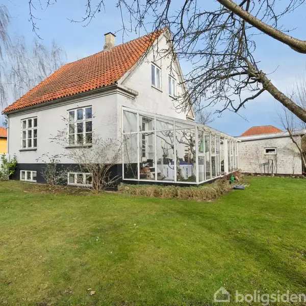 Et hvidt hus med rødt tag står stille omgivet af en velholdt have. En glaseret udestue er knyttet til huset. Omkringliggende træer uden løv ses mod en blå himmel.