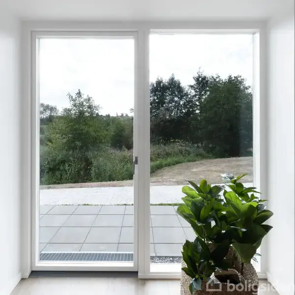 Et stort glasdørsparti står åbent mod en terrasse. En grøn plante i en kurv står indendørs ved siden af døren. Udenfor ses en flisebelagt terrasse og træer i baggrunden.