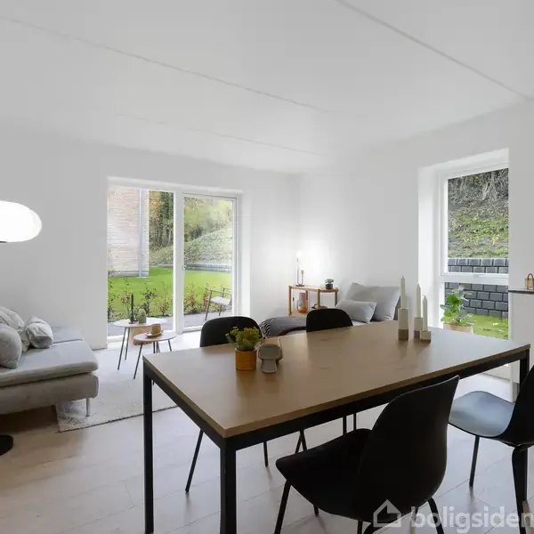 En spisestue indeholder et bord med stole og dekoreret med planter. I baggrunden er der en sofa ved siden af glasdøre, som fører ud til en grøn have.