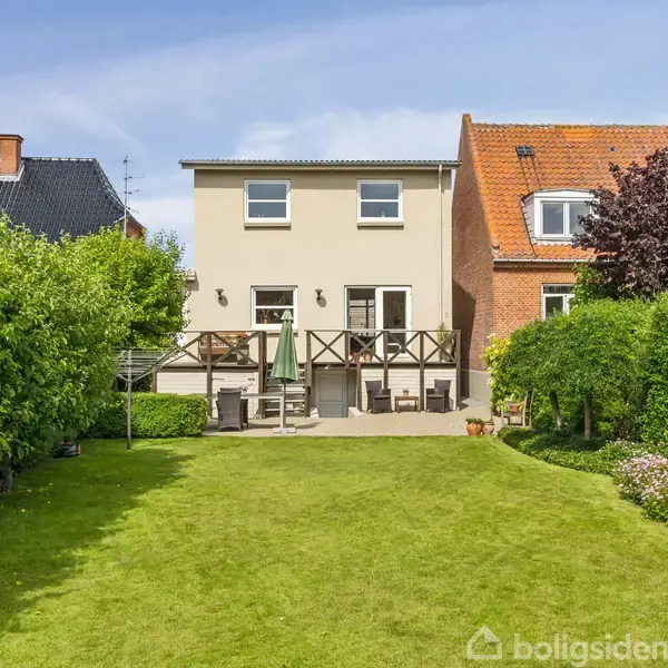 Et to-etagers hus med beige facade står forenden af en velplejet have med græsplæne og frodige buske. En terrasse har havemøbler og en parasol.