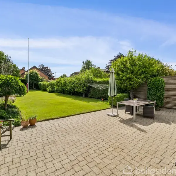 Terrasse med bord og parasol står på flisebelagt område. En frodig have omringet af buske strækker sig ud bagved med græsplæne og træer. Solrigt vejr. Ingen tekst fundet.