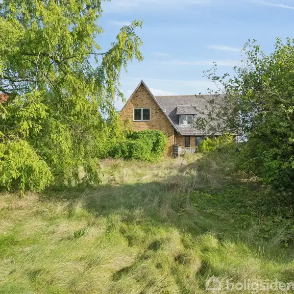 Et hus med spids tag står blandt højt græs og buske, omgivet af grønne træer under en blå himmel.