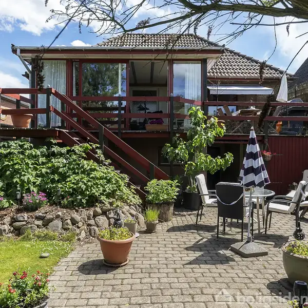 Et hus med en stor baghave, hvor der står en terrasse. Havemøbler og parasoller er placeret på fliserne i solen. Bede med planter omgiver området.
