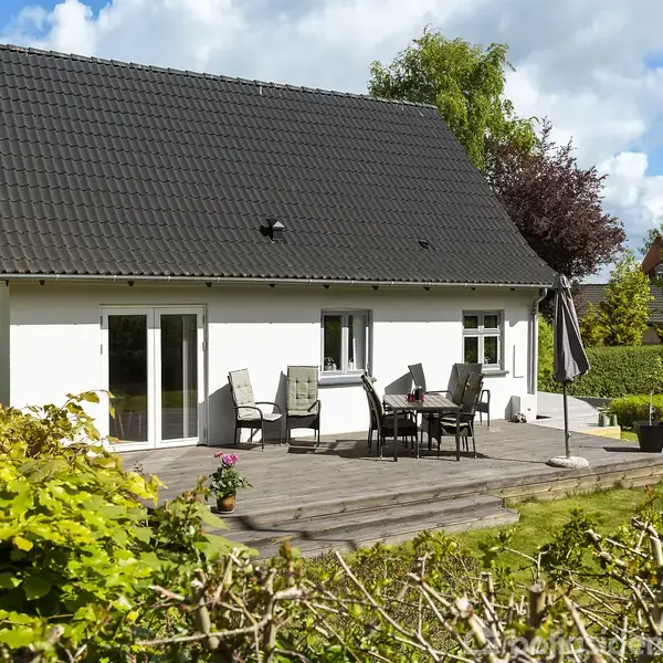 Et hvidt hus med sort tag står foran en veranda med havemøbler og parasol. Omgivet af velplejede buske og græsplæne under en blå himmel med skyer.