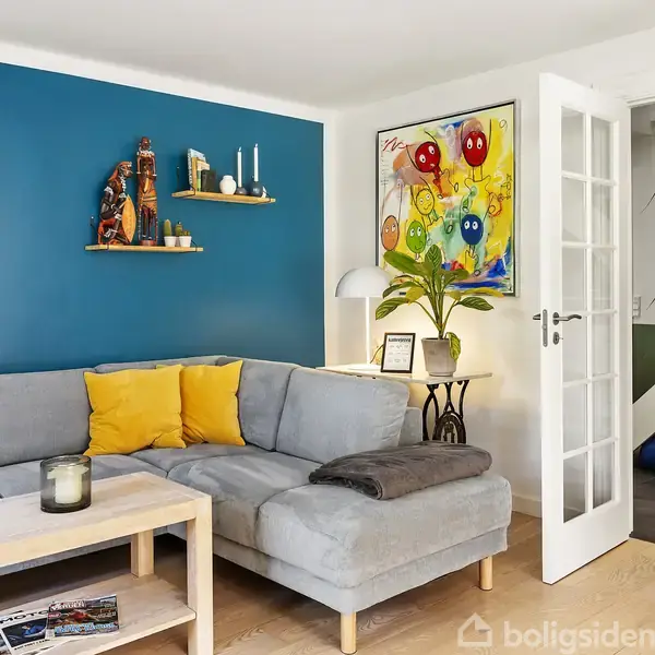 Gråt hjørnesofa med gule puder står mod en blå væg med hylde og verdenskort. Små borde og grøn plante er ved siden af, mens dør fører til næste rum.