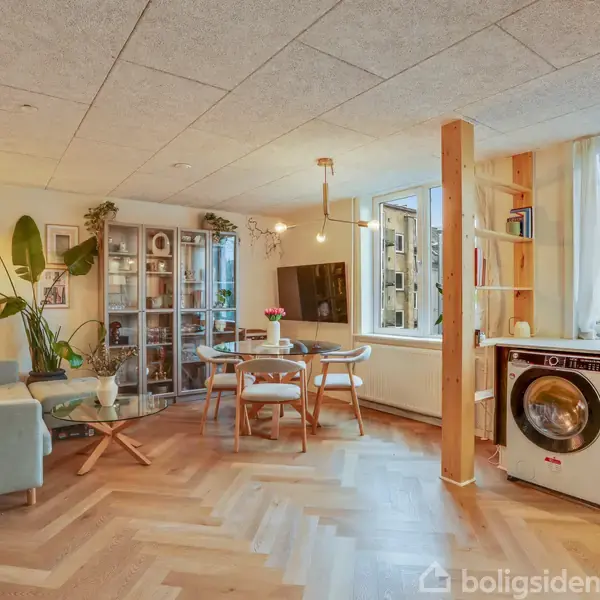 Stue med moderne indretning har sofa, spisebord og reol. Vaskemaskine står ved siden af køkkenet med udsigt til bygninger udenfor. En plante dekorerer hjørnet.