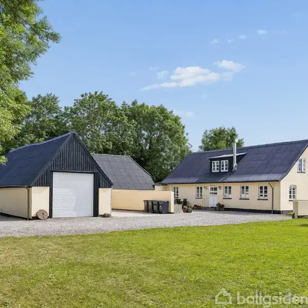 Et gult hus med mørkt tag og tilhørende garage sidder i en grøn have med grusvej. Træer omkranser området, og himlen er klar med nogle få skyer.