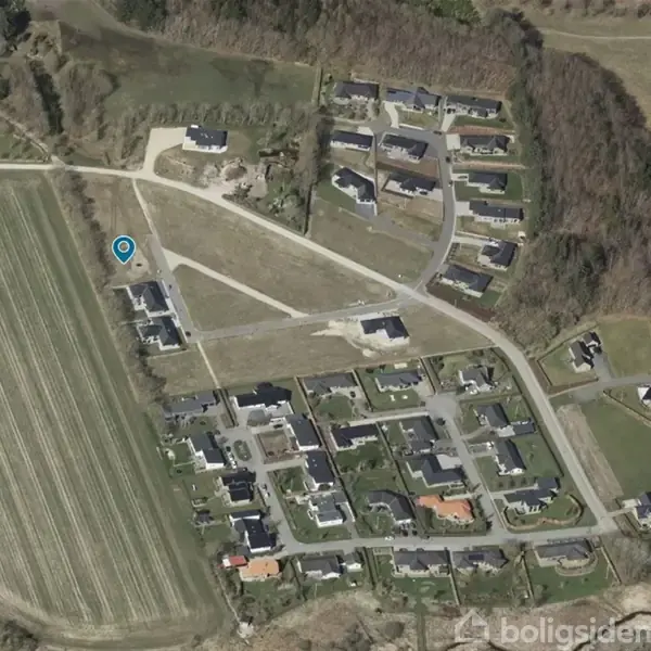 Luftfoto af et boligområde med huse og veje omgivet af skov og marker. En blå positioneringsmarkør viser en specifik placering blandt husene.