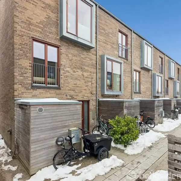 Rækkehus i mursten, omgivet af cykler og sne, står stille i et rækkehusområde. Flere tilbyggede skure langs bygningen. Klar blå himmel over det velordnede kvarter.