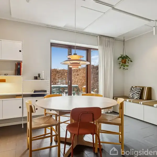 Rundt spisebord med træstole står i en lys spisestue. En moderne loftslampe hænger over bordet. Væghængte hylder og et vindue med udsigt til have supplerer rummet.