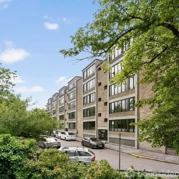 Spisebord med sorte stole står på et tæppe, omgivet af hvide vægge med kunst. Store vinduer og en dør åbner ud mod en terrasse med træer i baggrunden.