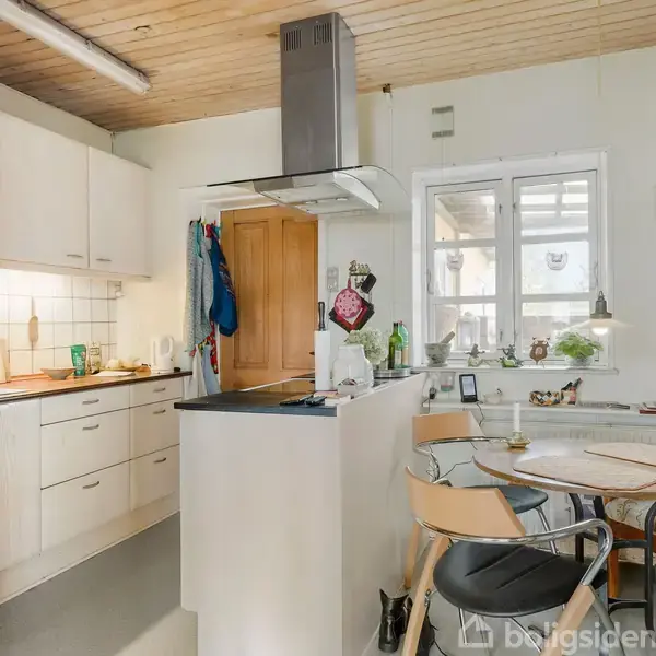 Køkken med lyse skabe og en kogeø, omgivet af en spiseplads med et rundt bord og stole. I baggrunden er der et vindue og en væg med en ur.