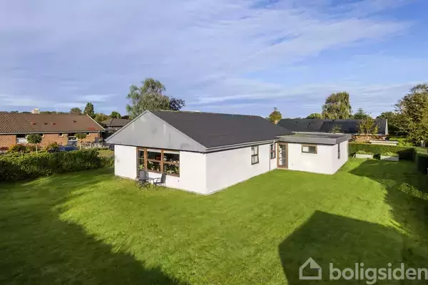 Huset har en hvid facade med sorte tage og store vinduer. Det står i en velplejet have omkranset af hække. I baggrunden ses flere huse og træer under en blå himmel.