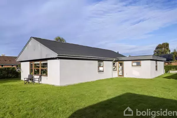 En enkeltstående grå bungalow står på en grøn græsplæne med et bord og to stole foran. Huset har store vinduer, og himlen er blå med enkelte skyer.