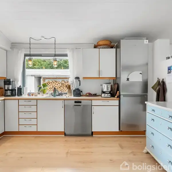 Et lyst køkken med hvide skabe og trækanter, indeholder opvaskemaskine, komfur og køleskab. Et lille bord står til højre, og vinduet viser en bunke brænde udenfor.