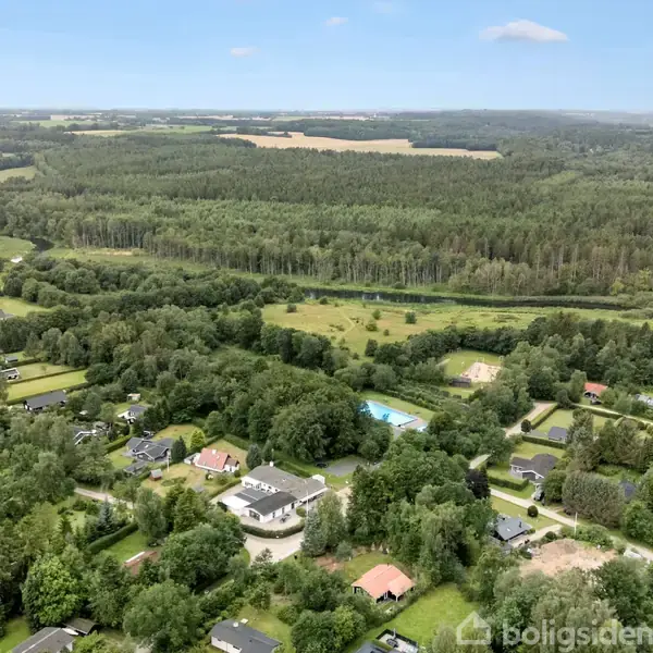Luftfoto viser landsby med huse indlejret i frodigt, grønt landskab. Skove og marker strækker sig mod horisonten, og en lys himmel huser enkelte skyer.