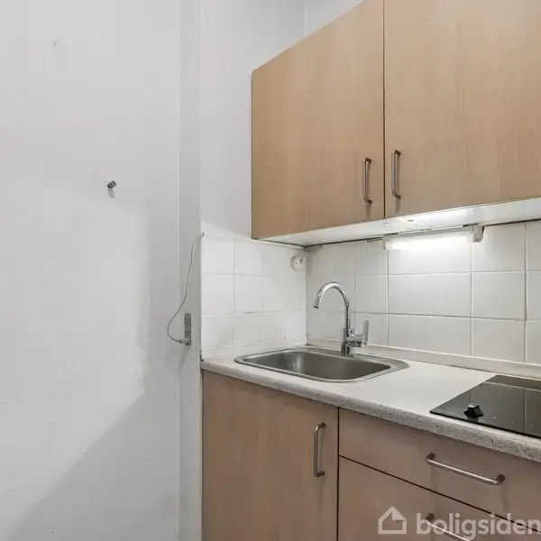 Køkkenområde med træskabe monteret på væggen. Håndvask og kogeplader er indbygget i en hvid bordplade. Flisevæg og belysning under skabene skaber et praktisk arbejdsområde.