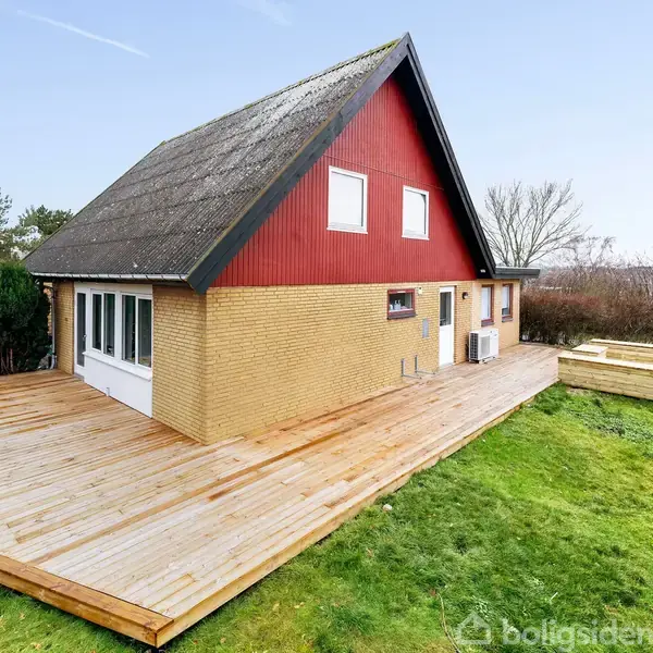 Hus med spidst tag står på en træterrasse. Det brune murværk og røde gavl tilføjer farve. Omgivet af en græsplæne, buskads og træer.