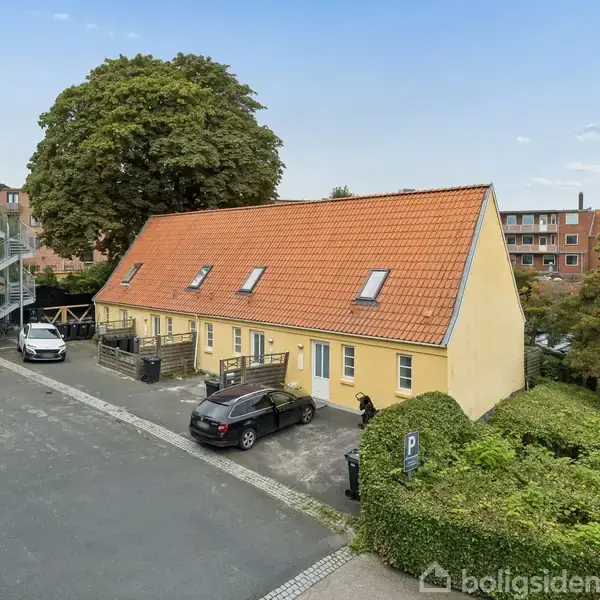 Et langt, gult rækkehus med rød tagbelægning står på en asfalteret parkeringsplads. Flere biler er parkeret foran, mens grønne buske omkranser området.
