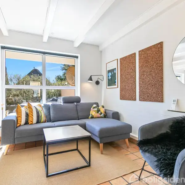 En stue med en mørkegrå sofa med farverige puder, placeret ved et stort vindue. Et sort bord foran sofaen. En grå lænestol til højre. Lyse fliser på gulvet.