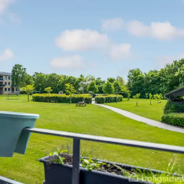 En altankasse med planter og en blå potte står på en altan. Udsigt over en græsplæne med stier, træer og et havepavillon. Bygninger ses i baggrunden.