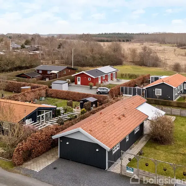 Flere huse med røde tage står i et parcelhuskvarter omgivet af hække og grønne græsplæner, der strækker sig mod åbent landskab i baggrunden.