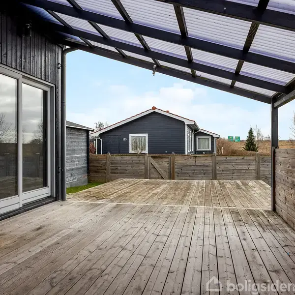 Overdækket terrasse med trædæk støder til sortmalet bygning. Store vinduer på venstre side. I baggrunden en anden sort bygning og træhegn, omgivet af åbent landskab og blå himmel.