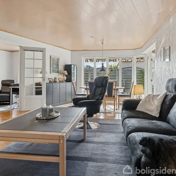 En stue med mørk sofa og lænestol foran et sofabord. Rummet åbner mod spiseområde gennem glasdøre. Trægulve og loft, med lysindfald fra vinduer mod haven.