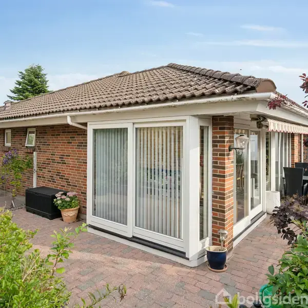 Et moderne murstenshus med store vinduer og et skråt tag står på en flisebelagt terrasse. Omgivet af grønne planter og en have, viser huset en afslappet, velholdt atmosfære.