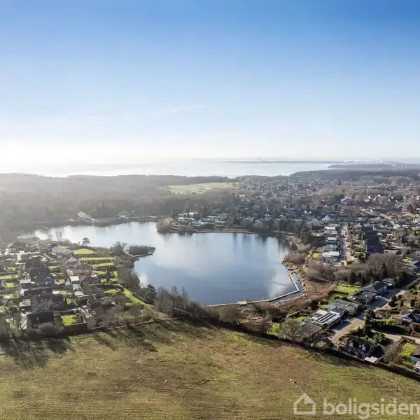 En sø ligger i centrum, omgivet af huse og grønne områder i en by. Skovområder og åbne marker strækker sig i baggrunden under en klar, blå himmel.